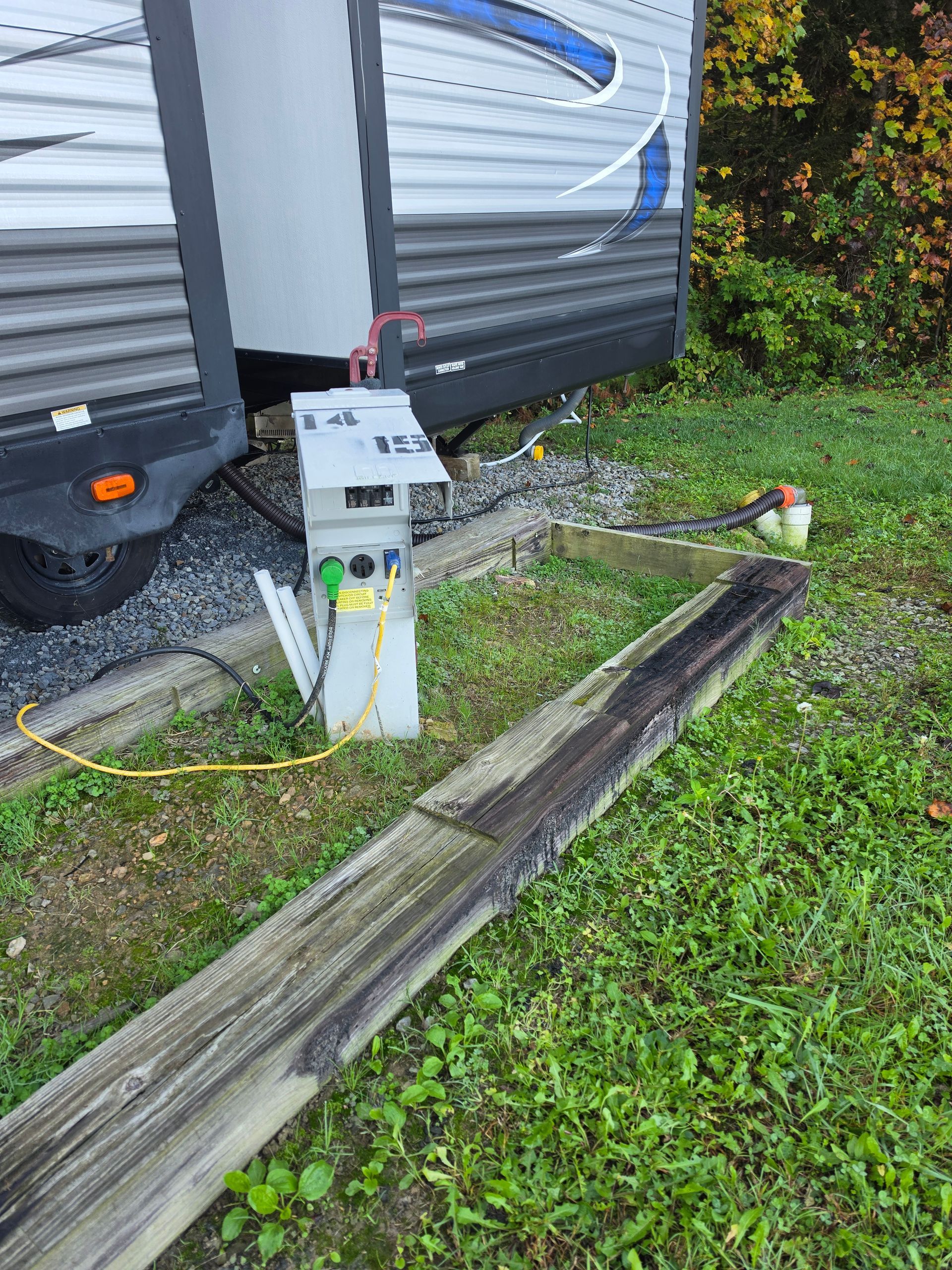 RV hookups beside a camper, with electrical pedestal and hoses on grass and gravel.