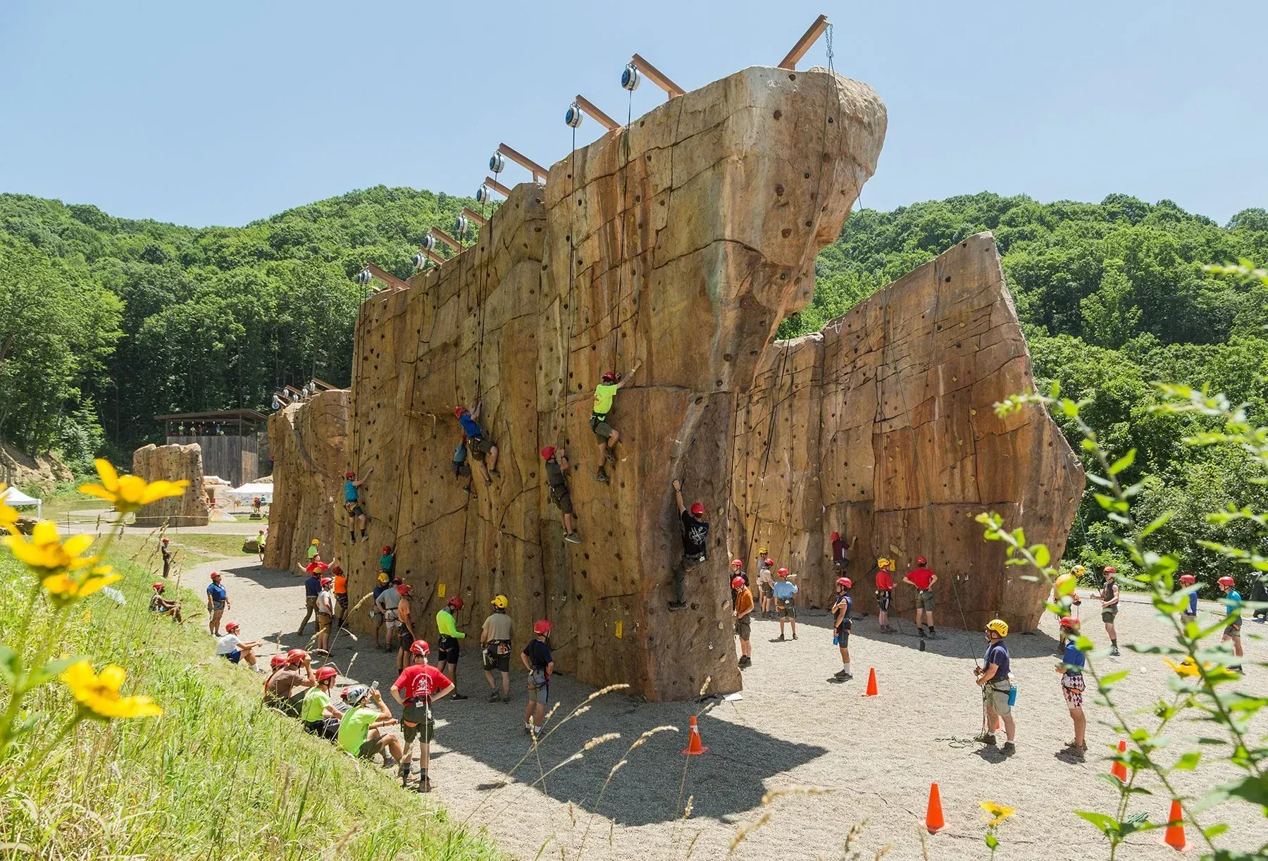 People climbing a tall outdoor rock wall at a training park, with cones and greenery around it
