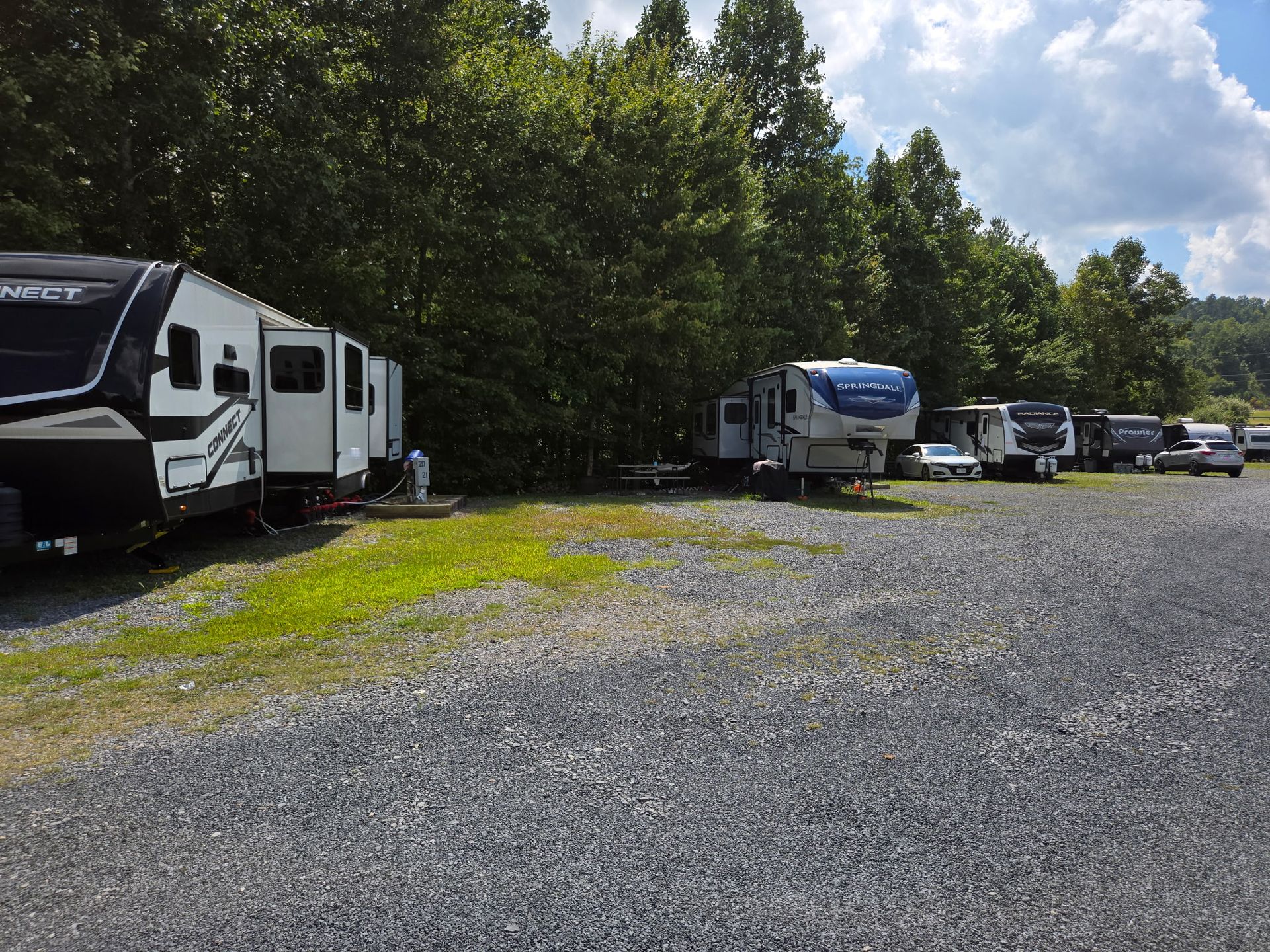 Row of RVs parked on a gravel lot beside a wooded area under a partly cloudy sky