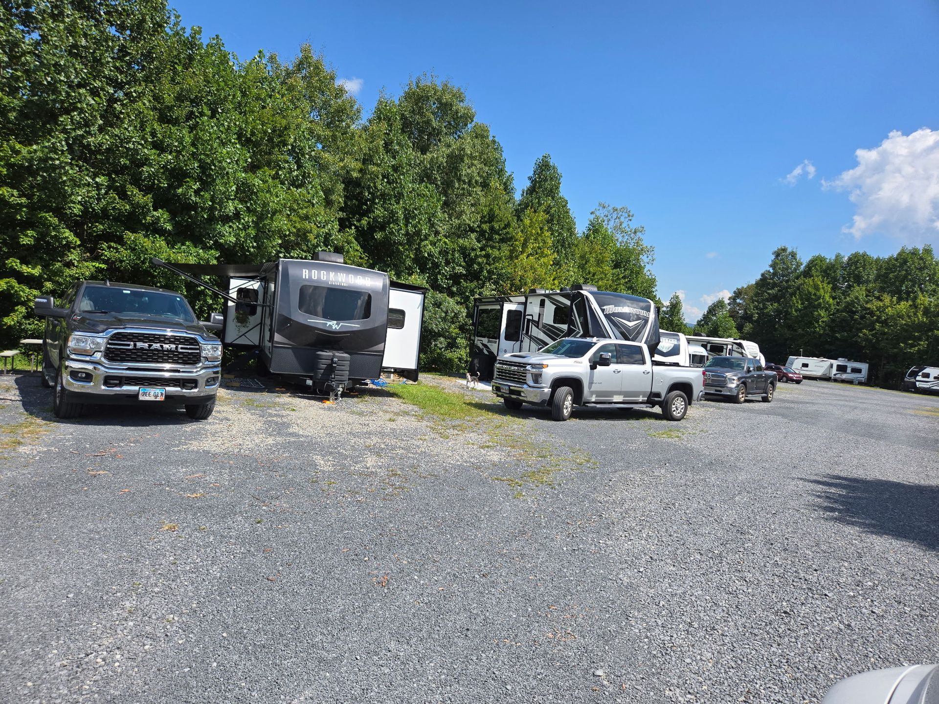 Parked trucks and RVs on a gravel lot beside trees under a blue sky