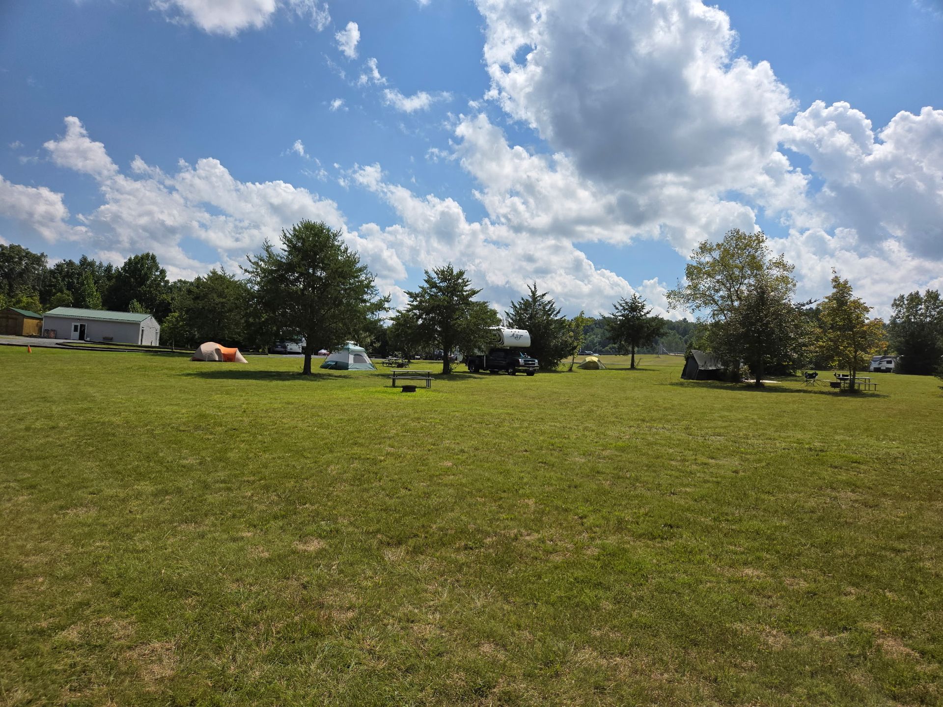 Grassy park with scattered trees and tents under a bright blue sky with white clouds
