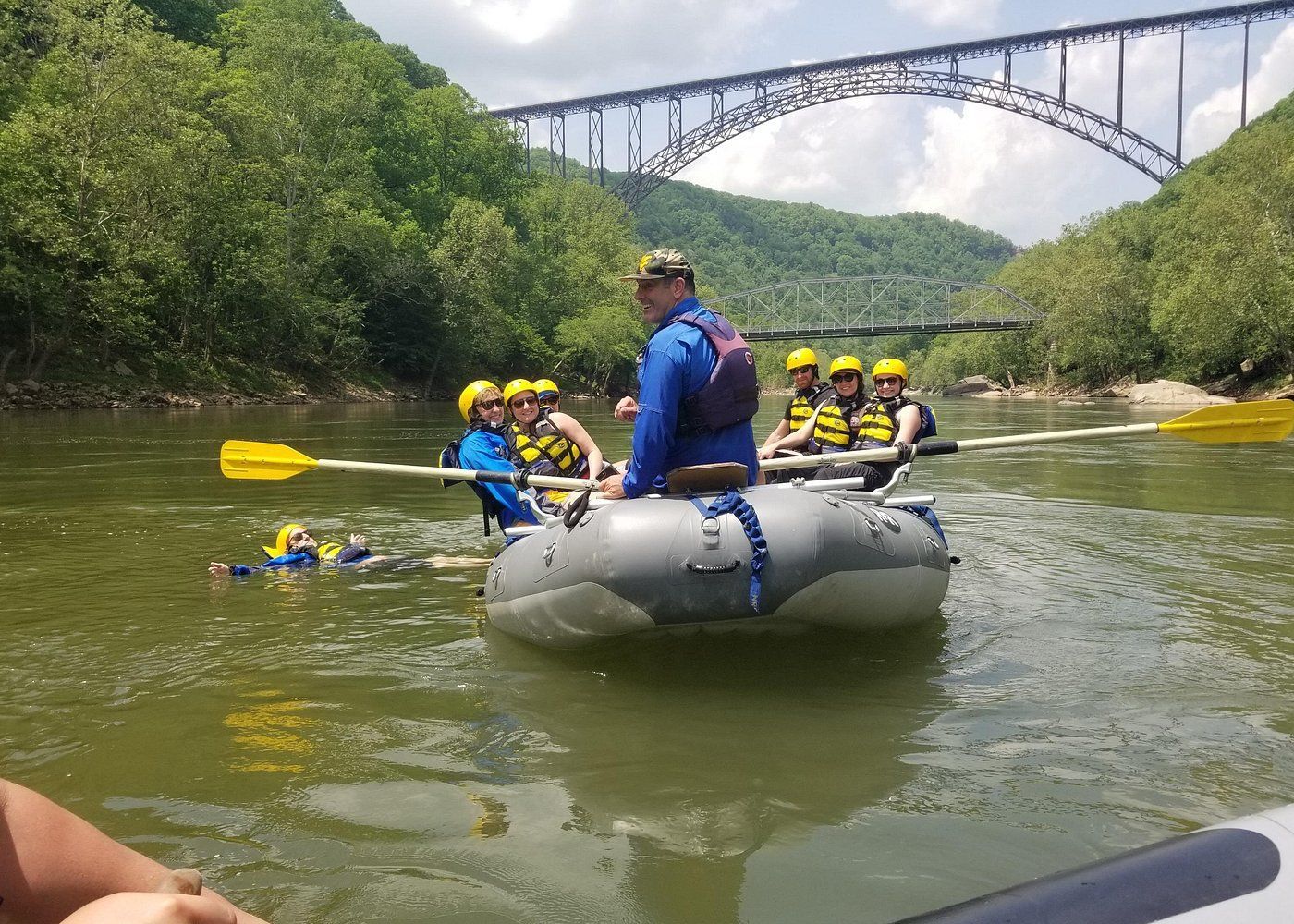 Rafting group in yellow helmets on a river beneath a tall bridge, with lush green hills around.