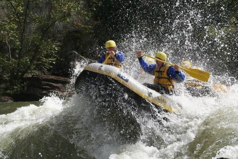 Two rafters in yellow helmets and life jackets ride a white inflatable boat through churning rapids