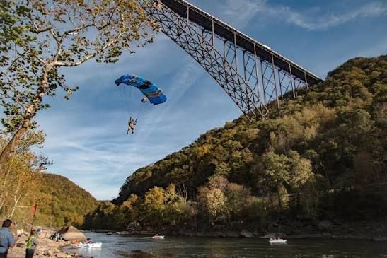 Skydivers descend near a large bridge over a river, with trees and hills lining the shore.