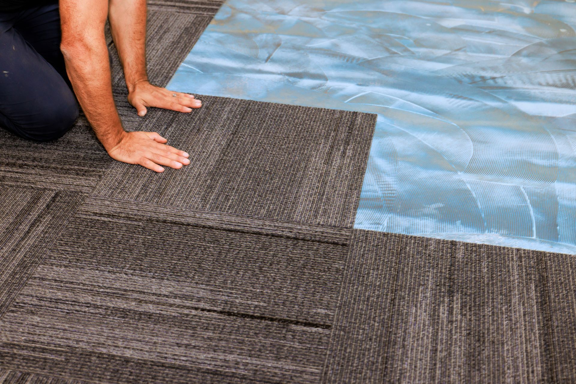 Person installing dark carpet tile, kneeling on installed tiles. Person installing dark carpet tile, kneeling on installed tiles.