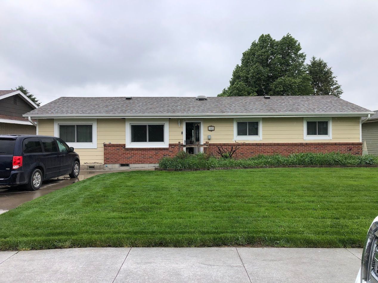 Yellow house with brick base and green lawn under cloudy sky. Dark minivan in the driveway.
