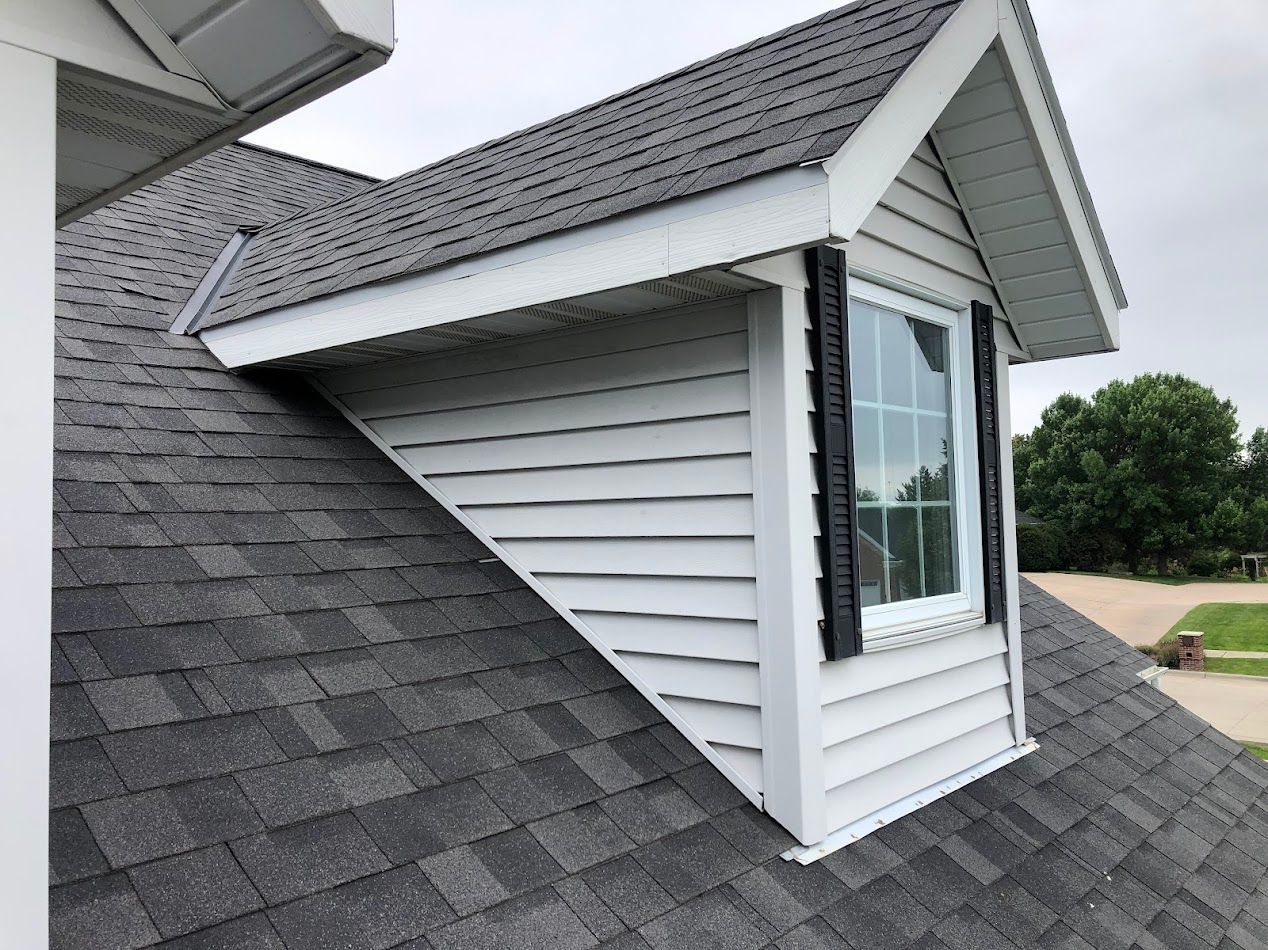 Dormer on a house roof with gray shingles, white siding, and a window with black shutters.