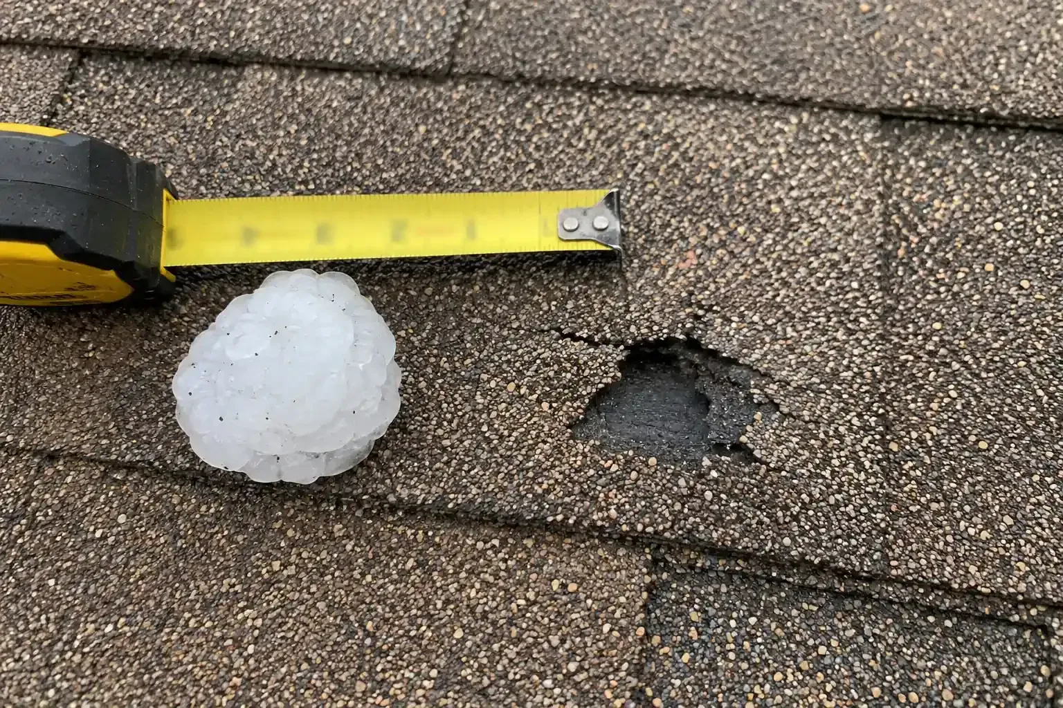 Large hailstone beside a tape measure on a roof with broken shingles