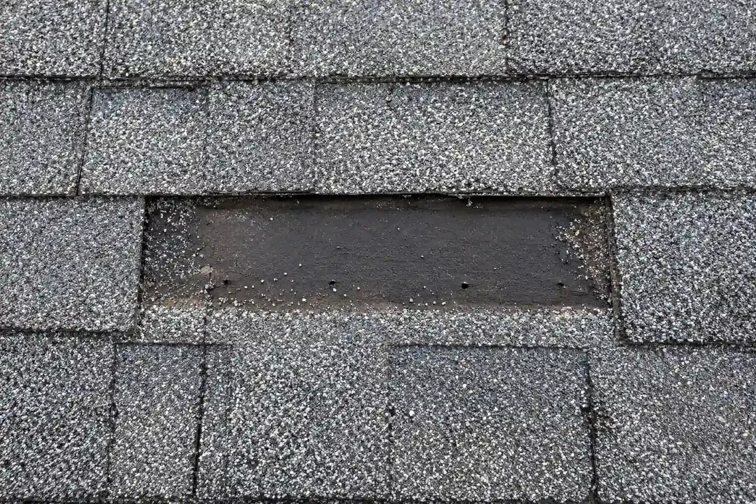 Bare spot on a shingle roof where wind tore away a shingle, showing the underlayment
