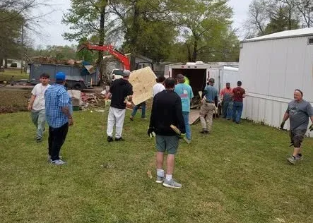 People standing in a grassy yard near trailers and an excavator, with debris and trees in the background.