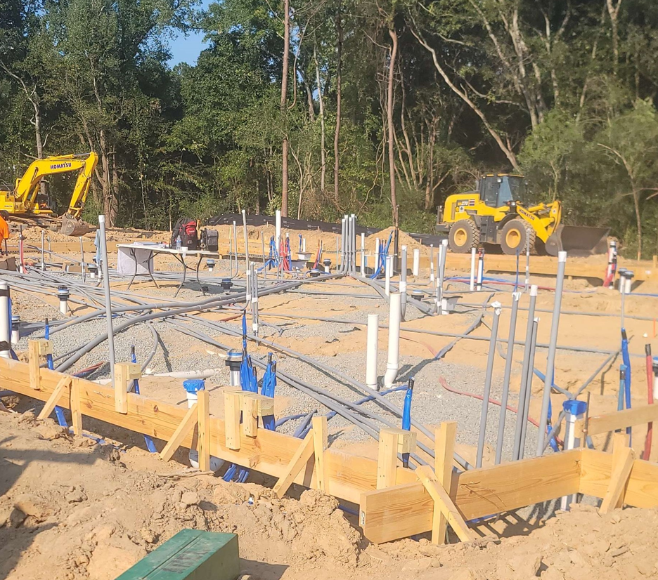 Construction site with concrete slab formwork, pipes, and yellow excavators in a wooded area