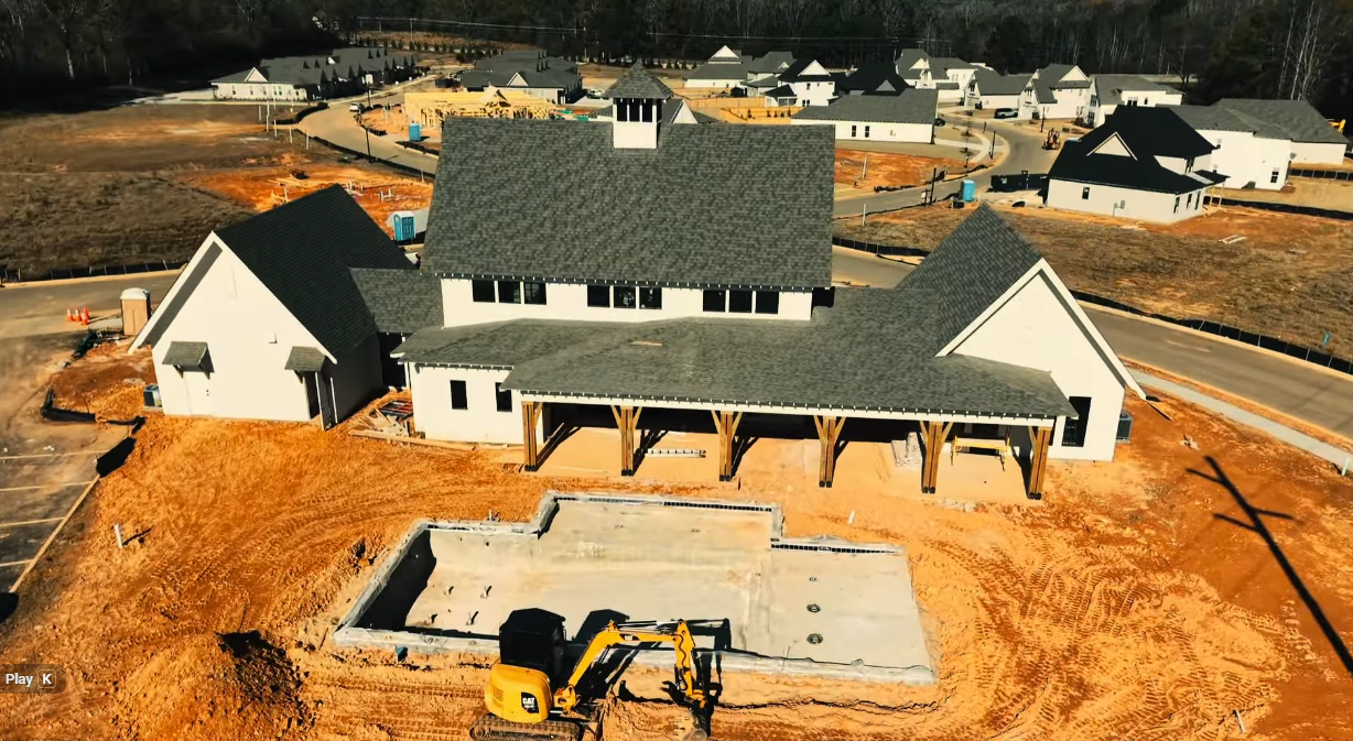New house under construction with a large roof, gravel yard, and a yellow excavator in front.