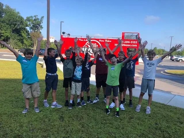 A group of young boys are standing in front of a red bus with their arms in the air.