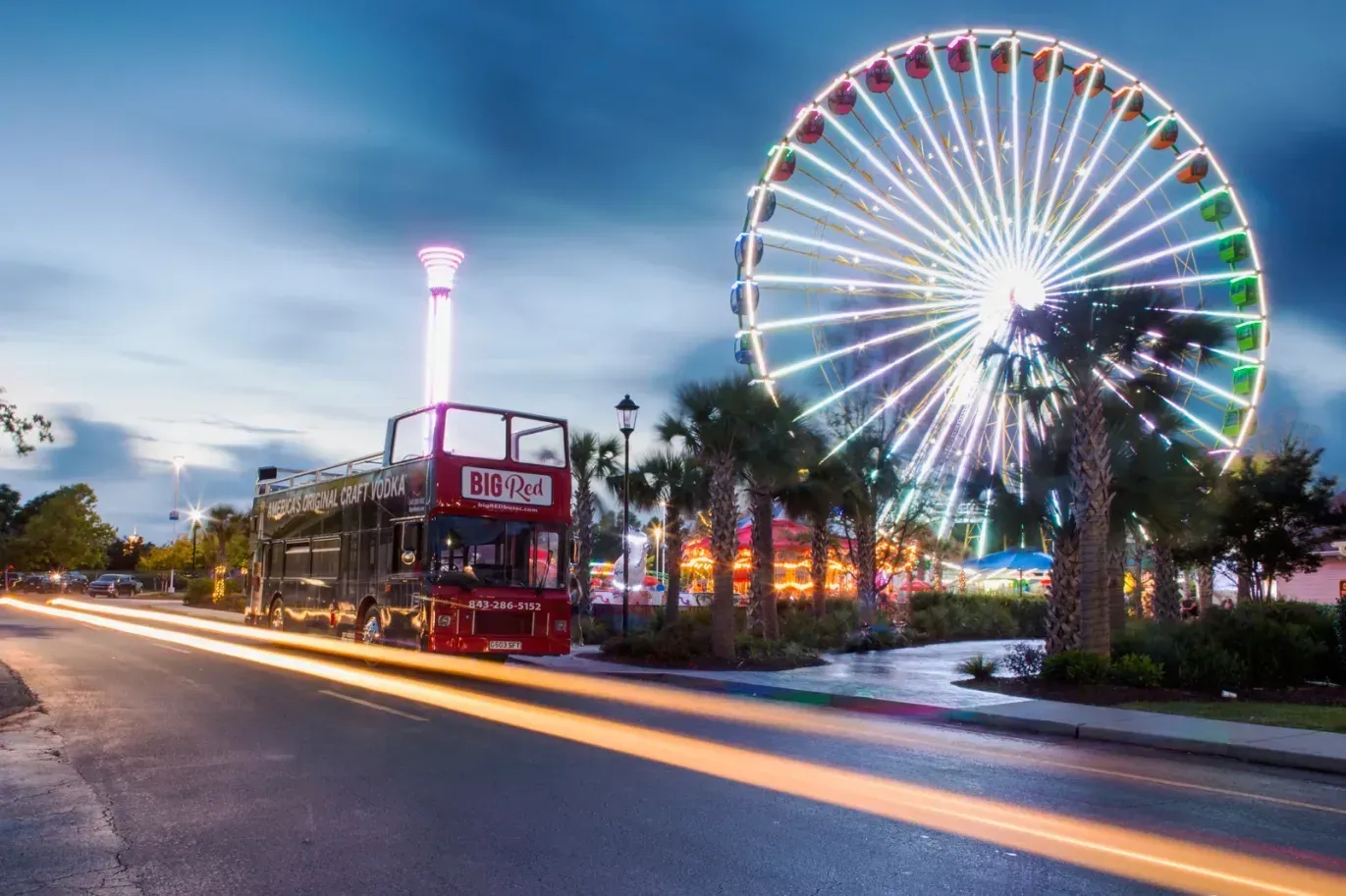 A double decker bus is driving down a street in front of a ferris wheel.