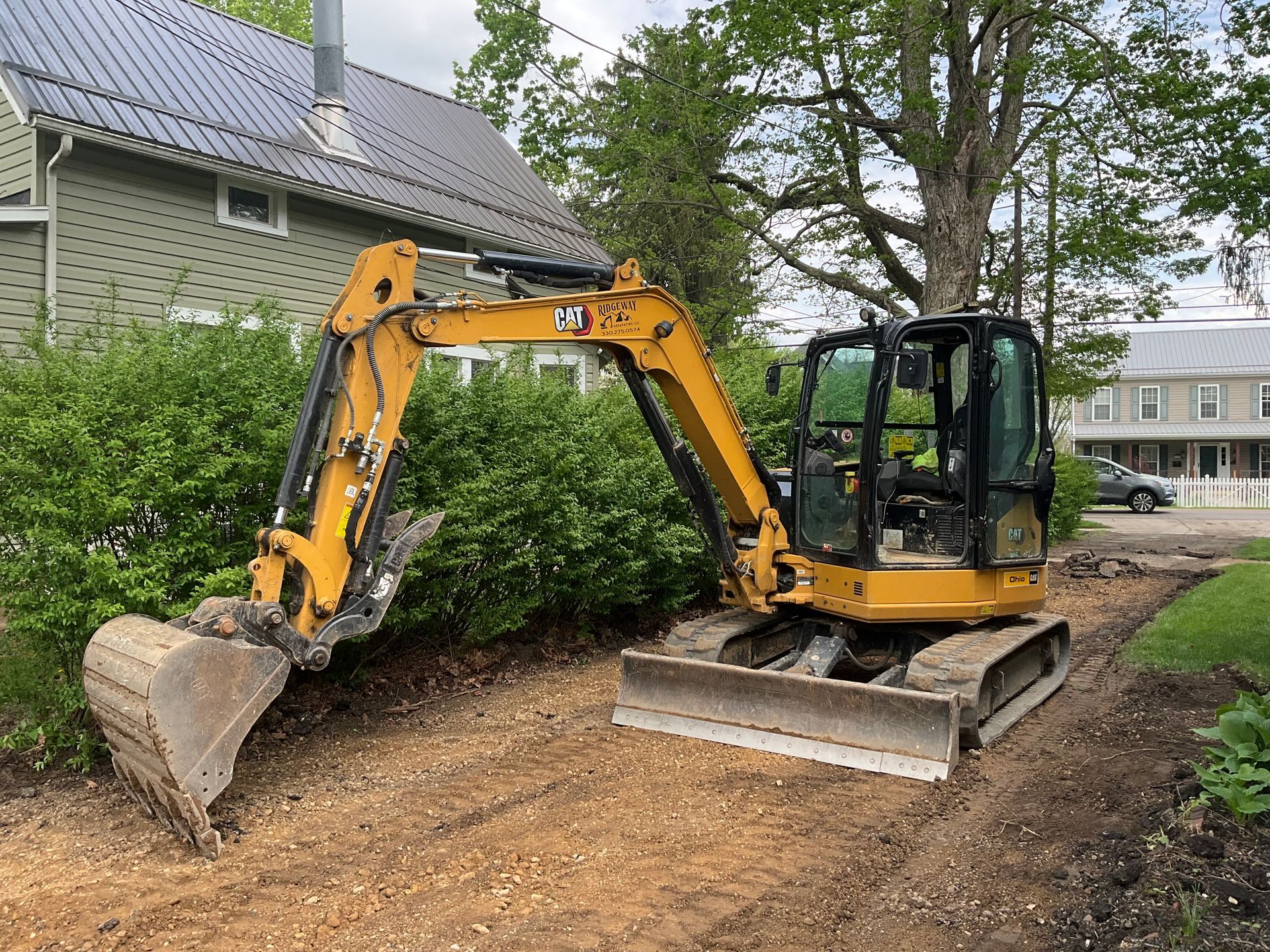 Yellow excavator clearing a dirt path beside a house and bushes