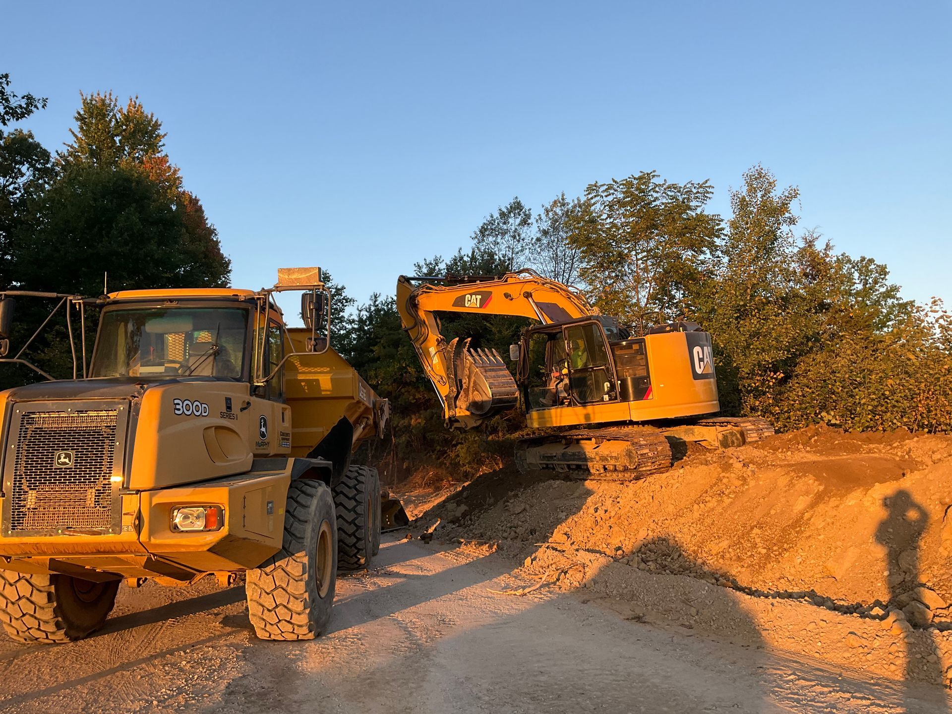Yellow construction truck and excavator moving dirt at a roadside work site in daylight