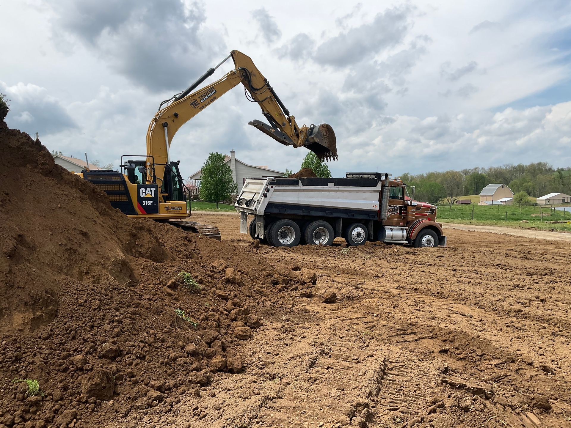Excavator loading dirt into a dump truck at a muddy construction site under cloudy skies
