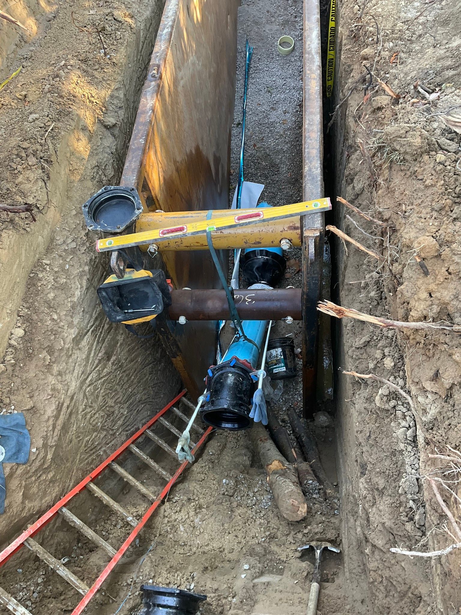 Workers at a deep trench with a ladder, yellow beam, and utility pipe repair equipment.