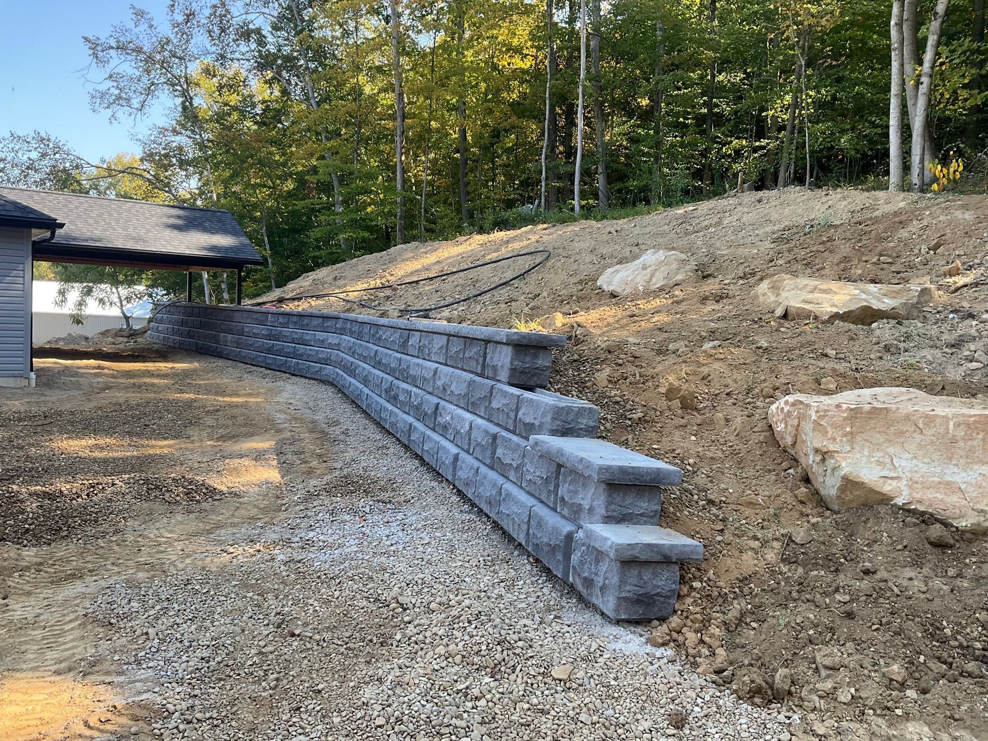 Retaining wall of gray stone blocks along a gravel driveway beside a wooded hillside