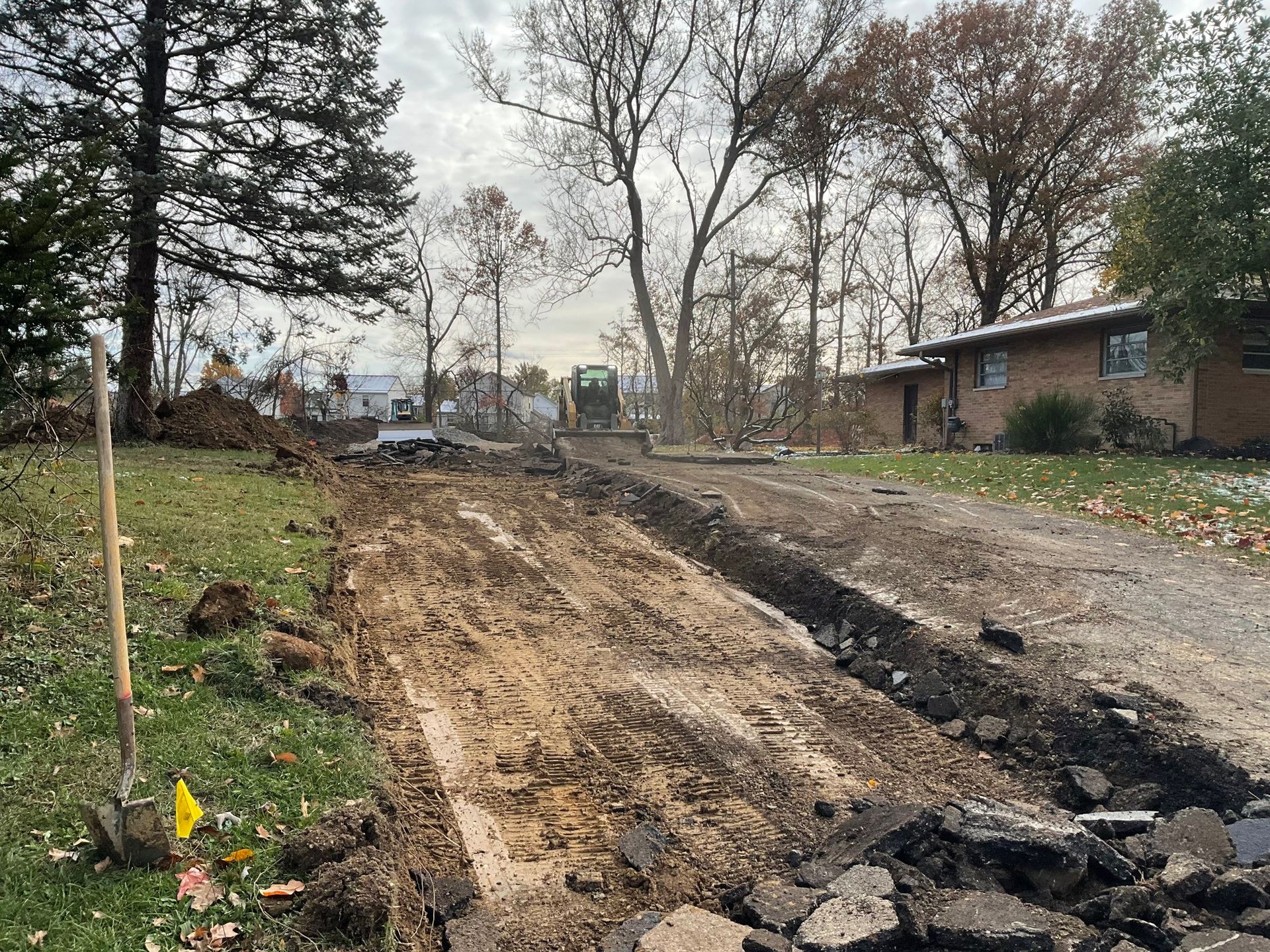 Partially dug-up dirt road beside houses and trees, with muddy soil piled along the side