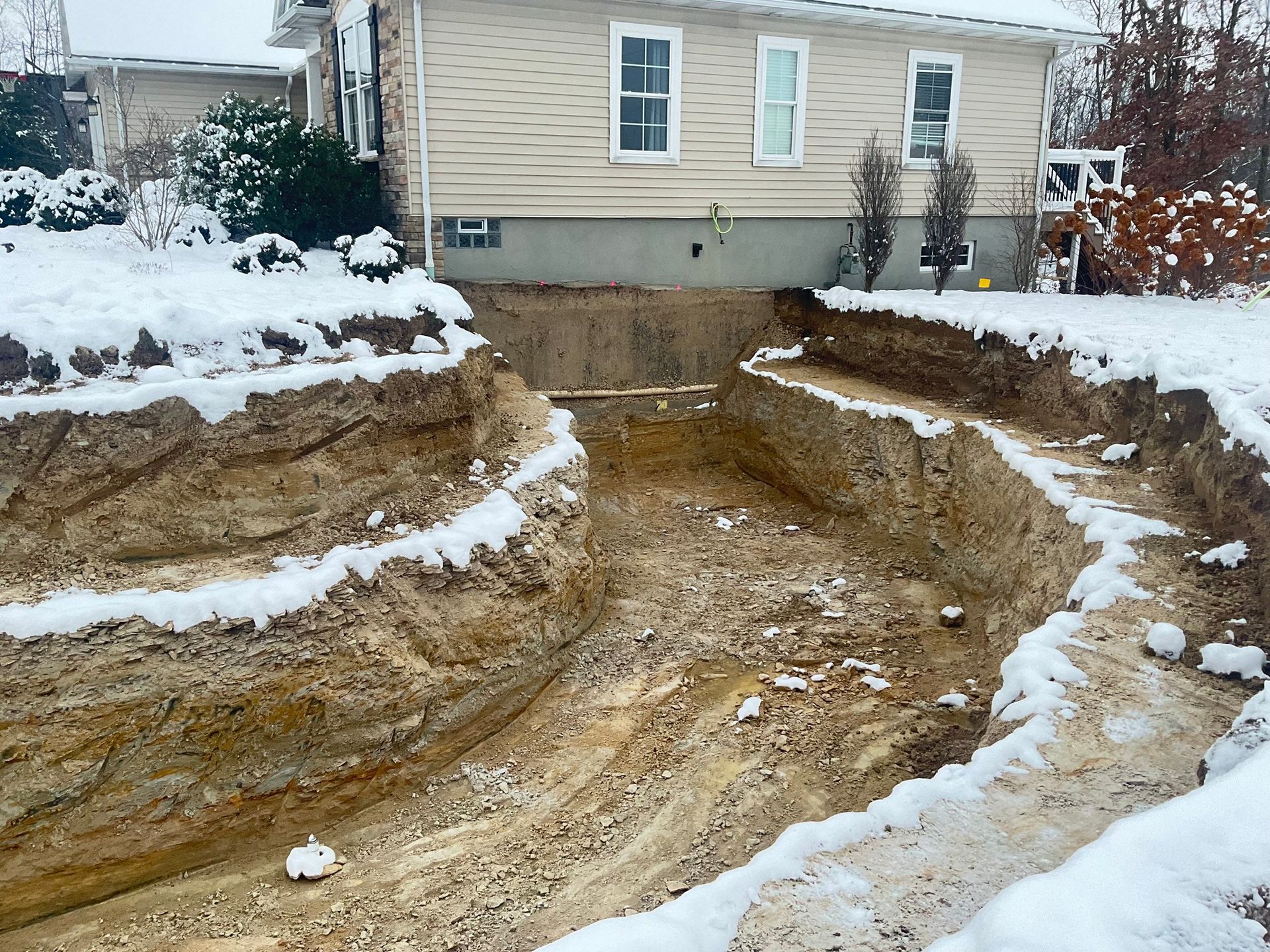 Snowy backyard excavation with a large dirt trench in front of a house