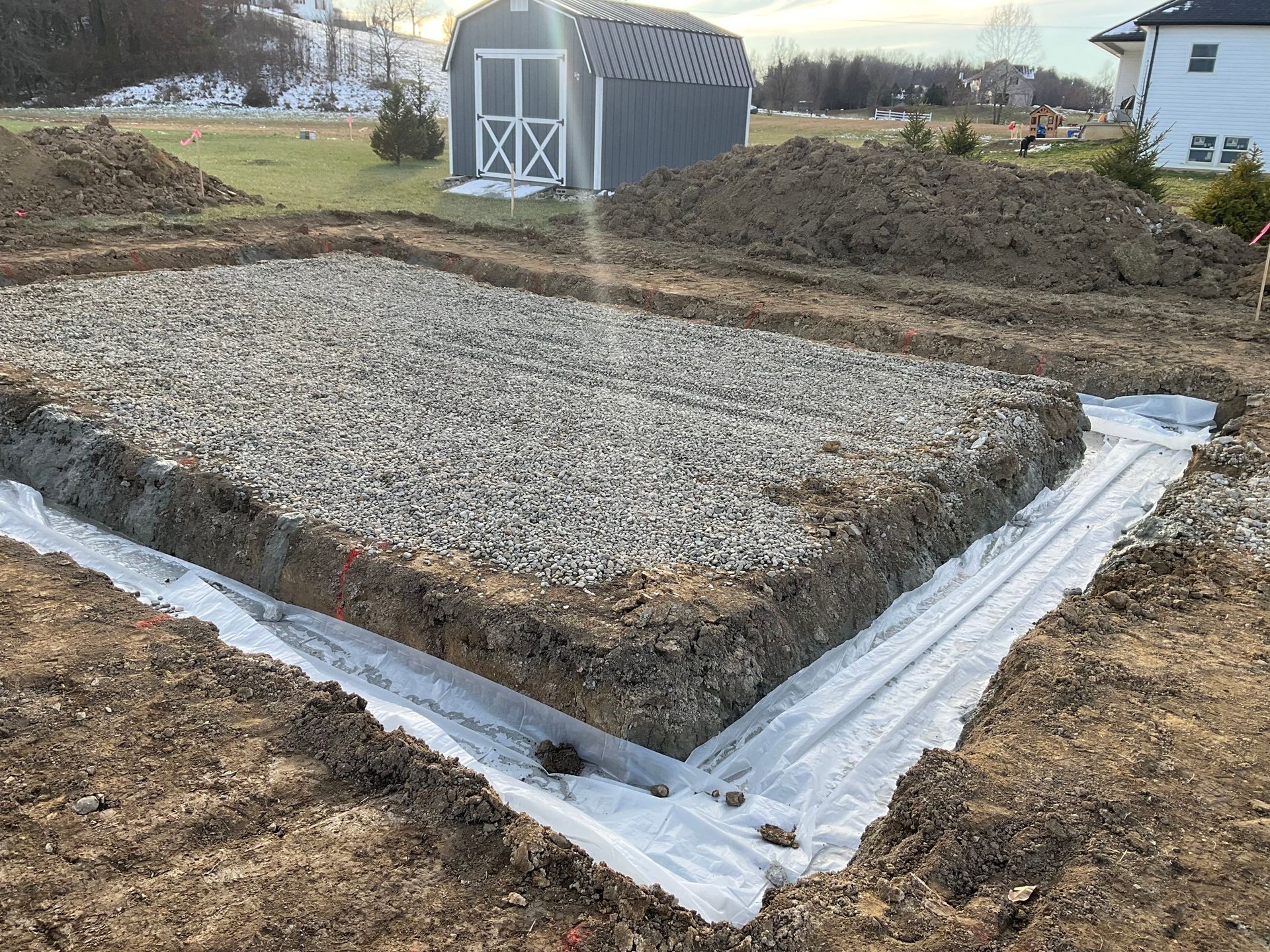 Gravel foundation pad bordered with white plastic sheeting beside a gray shed on a rural lot