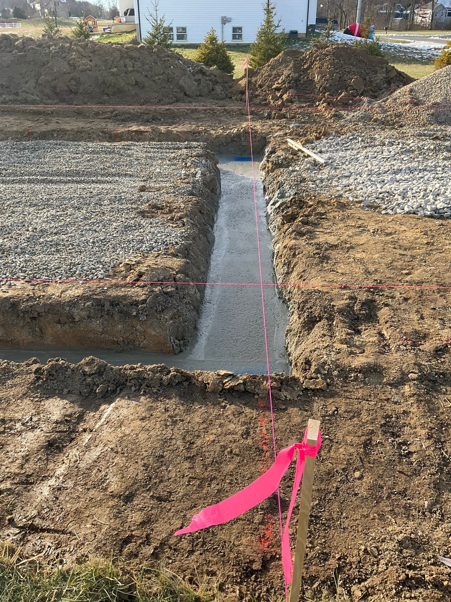 Construction site trench with gravel-filled sections and a narrow concrete channel marked by pink tape