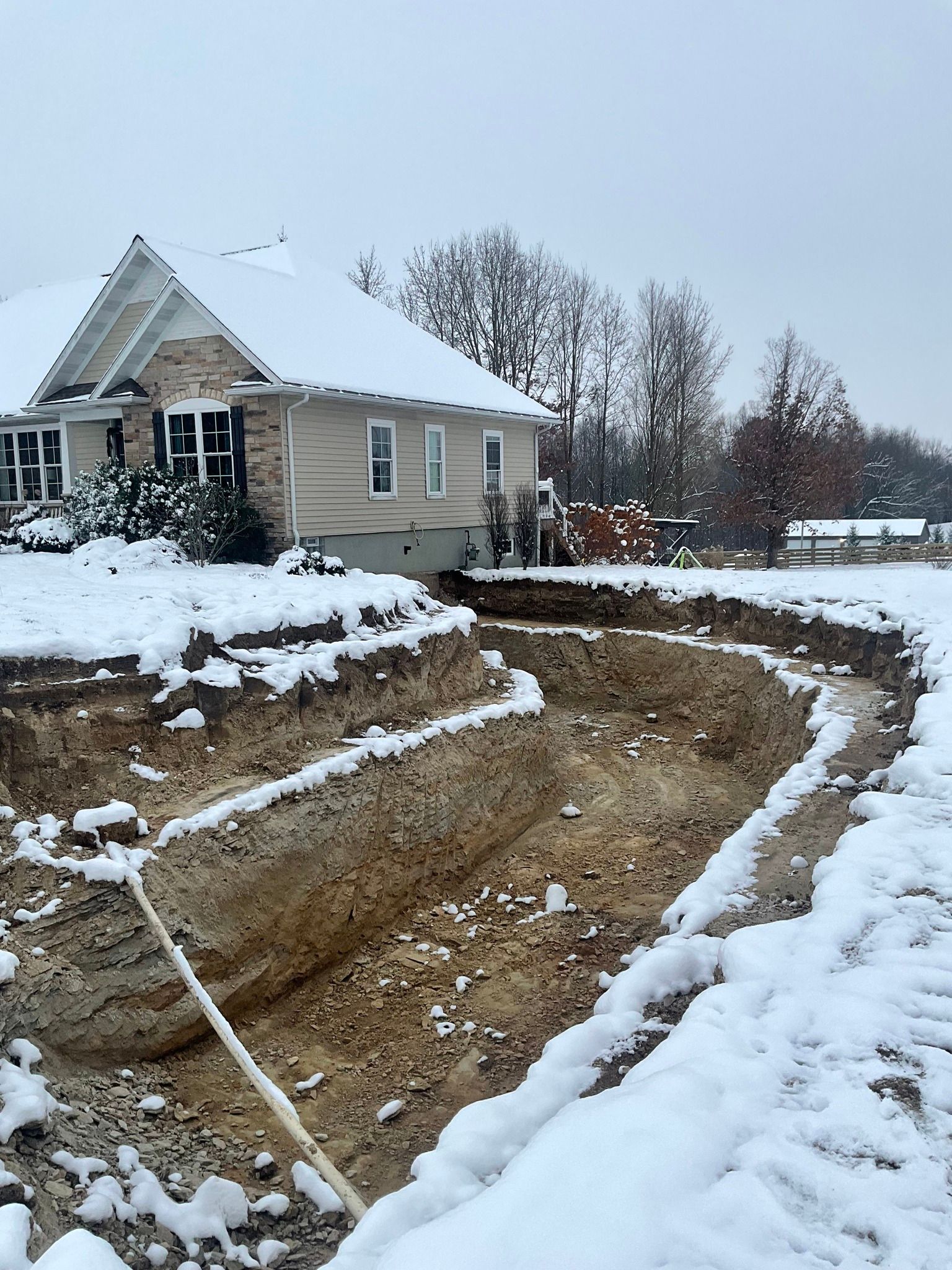 Snowy construction site beside a beige house with a dug-out yard and dirt paths