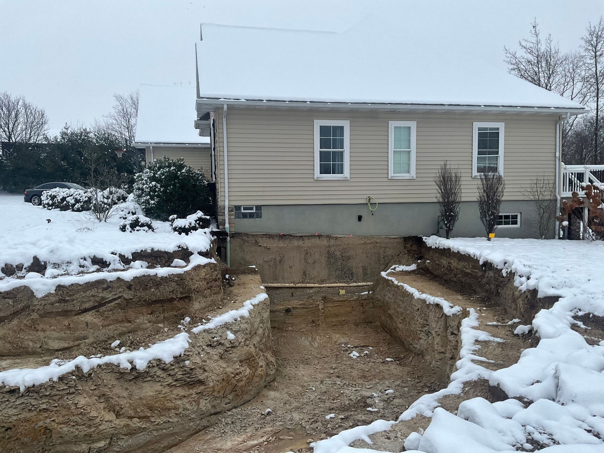 Snowy backyard with a house, showing a large excavated trench or foundation work in progress.