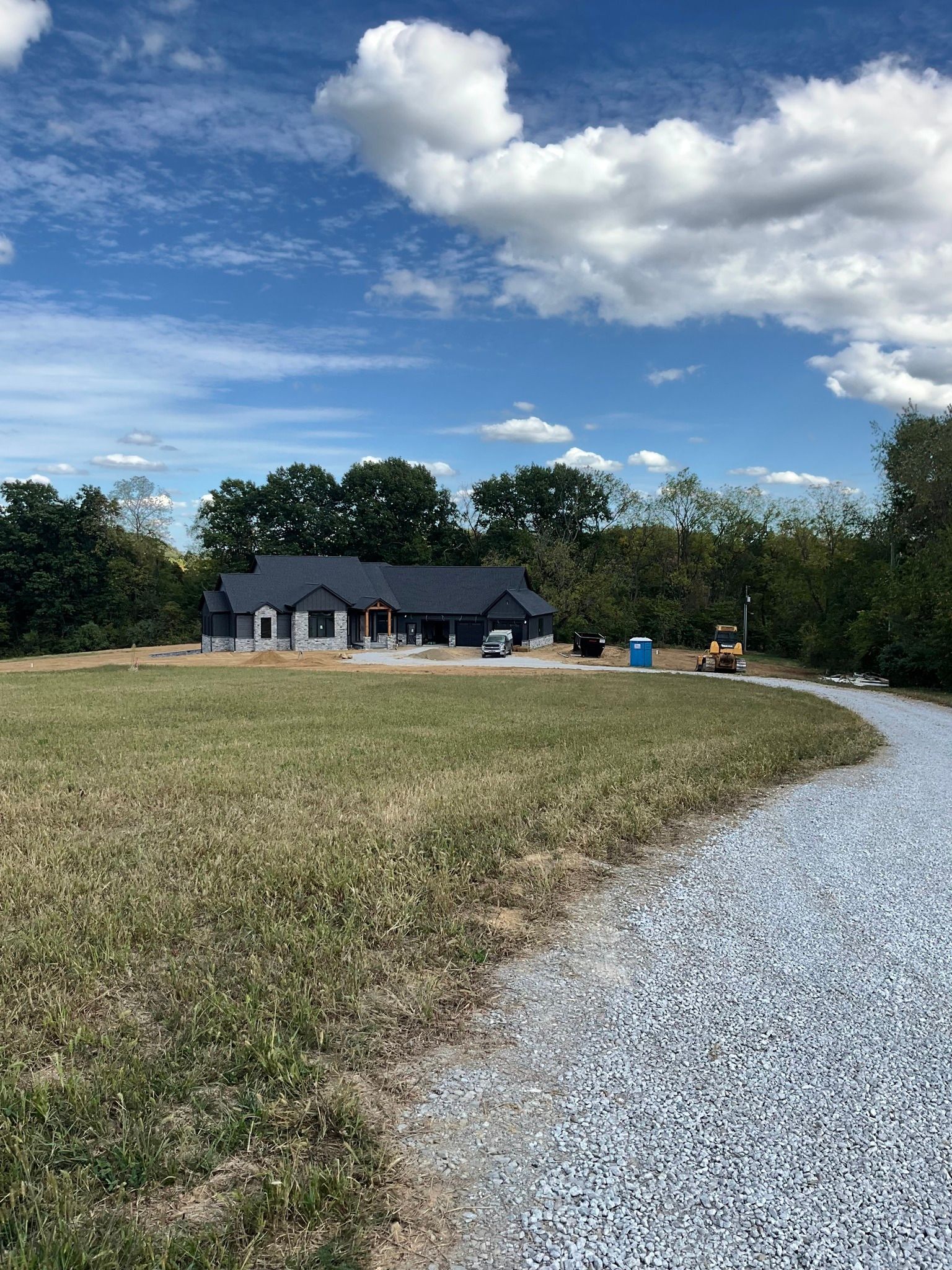 Gravel driveway curving toward a small dark house in a grassy field under a partly cloudy blue sky