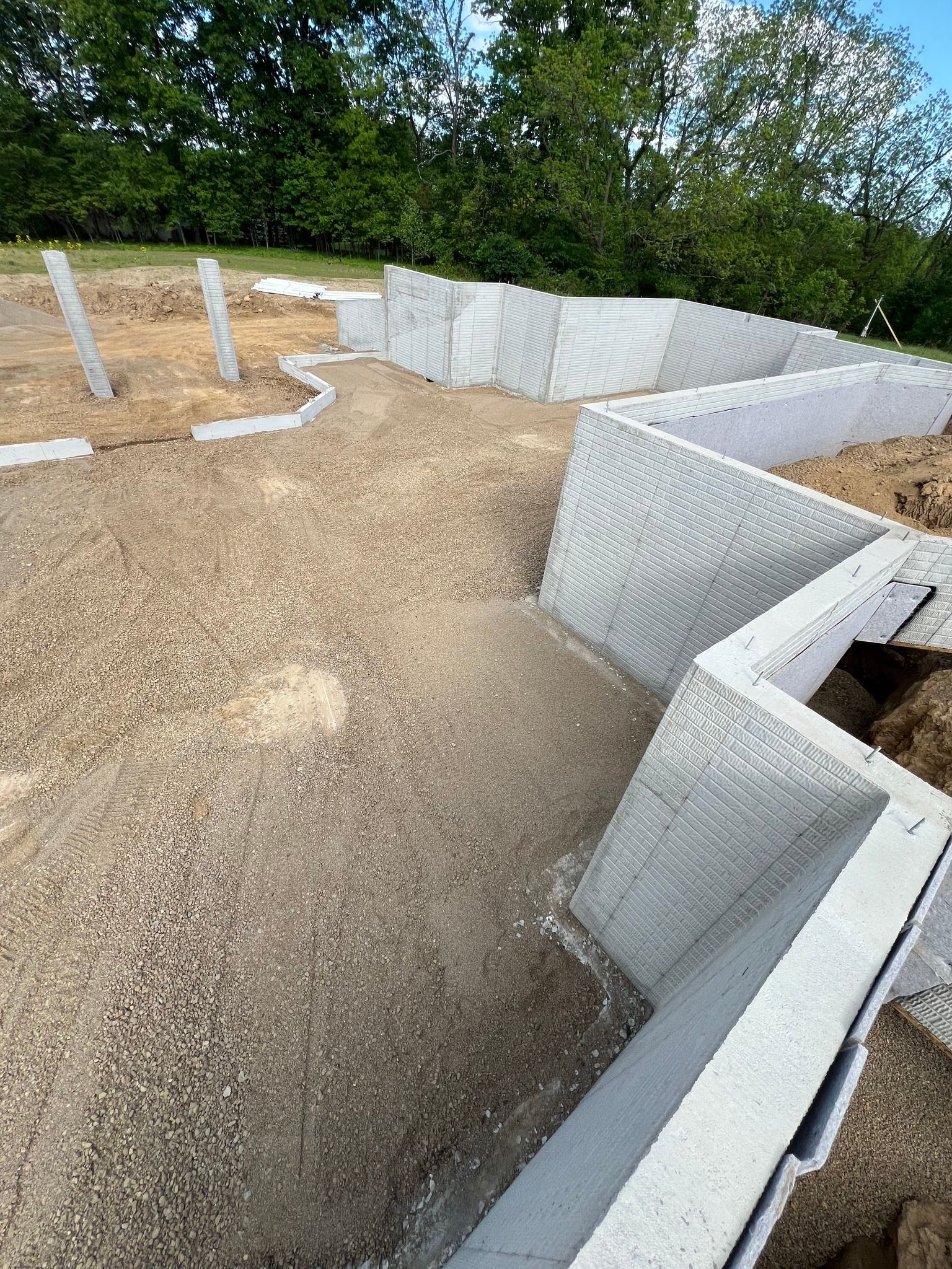 Gravel foundation area with white insulated concrete forms and rebar posts outdoors under trees
