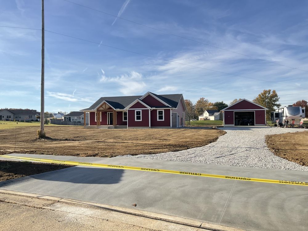 Red house and detached garage on a gravel driveway in a grassy, partly built residential lot.