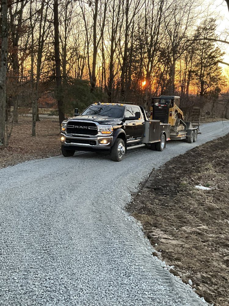 Tow truck hauling a black pickup on a gravel road at sunset through bare trees