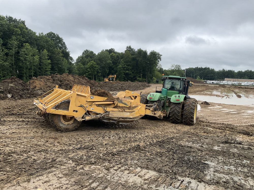 Green tractor towing a yellow earthmover across a muddy construction site under cloudy skies