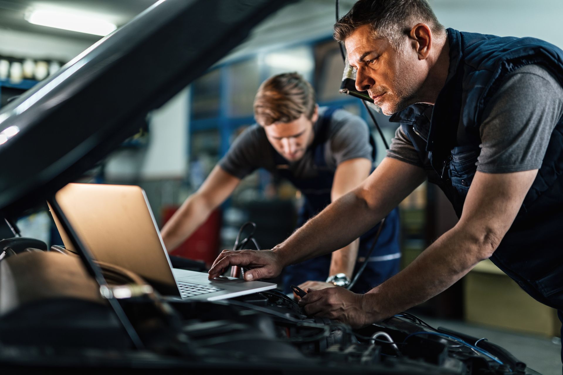 An auto mechanic is using a laptop while working on a car diagnostic with his coworker.