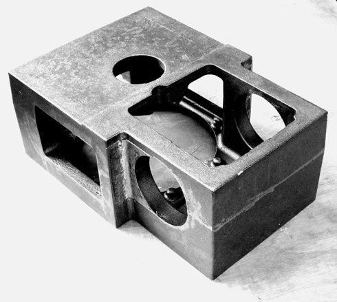 Un blocco di metallo grigio, lavorato a macchina, con diverse aperture rettangolari e circolari, su uno sfondo chiaro e uniforme.