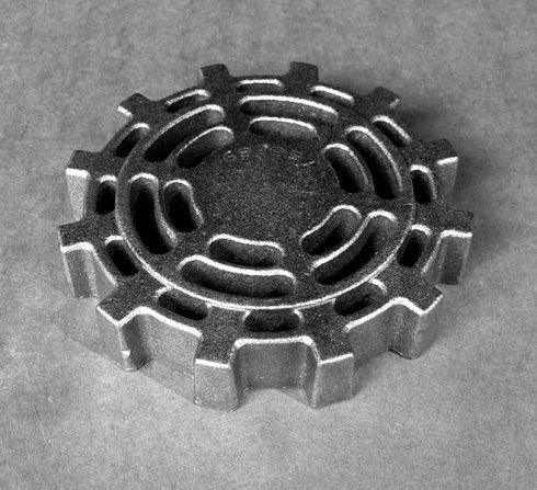 Un componente metallico circolare di colore argento con un bordo esterno a forma di ingranaggio e fessure di ventilazione decorate sulla sua superficie.