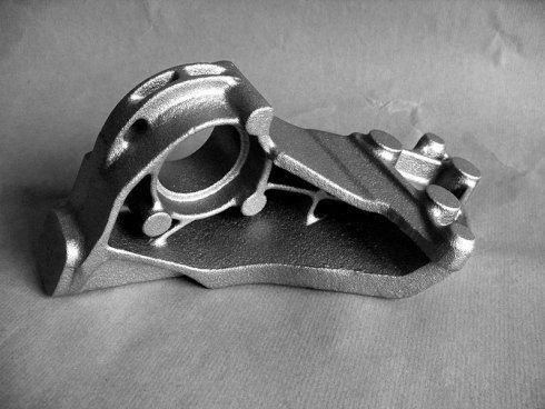 Componente meccanico in metallo pressofuso di colore grigio, in stile industriale, con apertura circolare centrale e punti di fissaggio tramite bulloni.