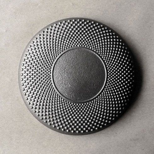 Un sottopentola circolare nero e testurizzato, con un centro liscio circondato da un motivo radiale di puntini metallici in rilievo.