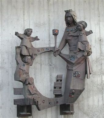 Una scultura a rilievo in metallo scuro e stilizzato, raffigurante una figura che tiene in braccio un bambino, collegata ad altre figure tramite una cornice circolare.