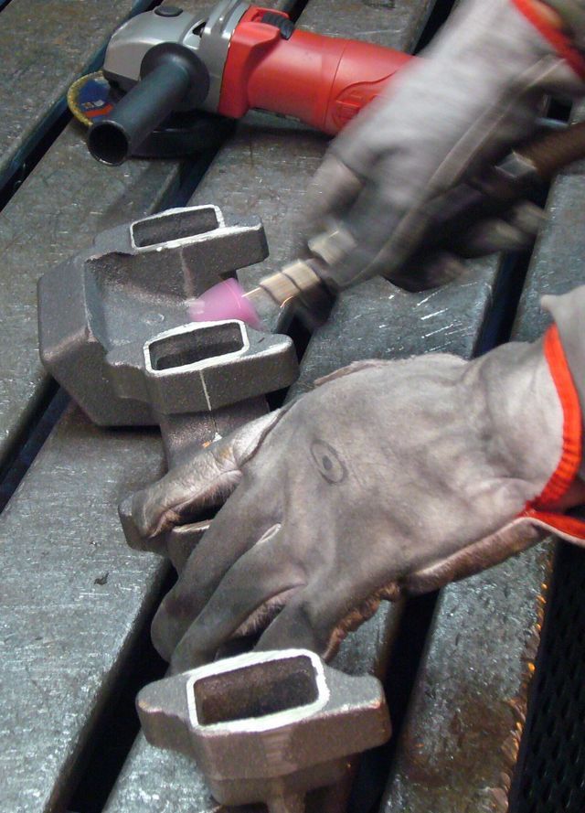 In un'officina, una persona con i guanti da lavoro utilizza un piccolo utensile abrasivo su un componente metallico.