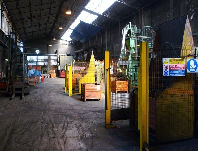 Vista interna di un ampio stabilimento industriale scarsamente illuminato, con macchinari pesanti, container e segnali di sicurezza.