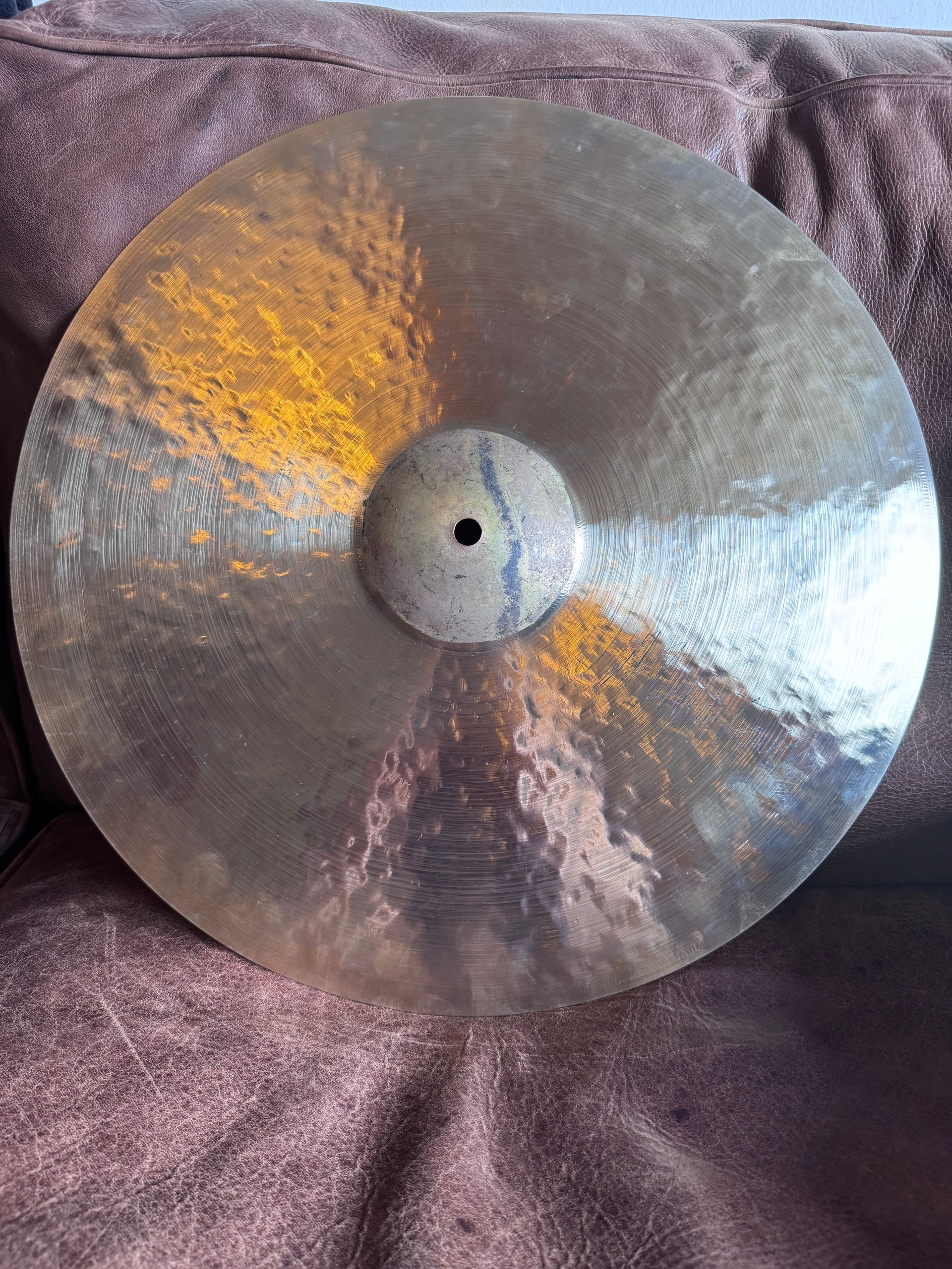 A tarnished, round bronze cymbal lies on a textured, brown surface. It has a central hole.