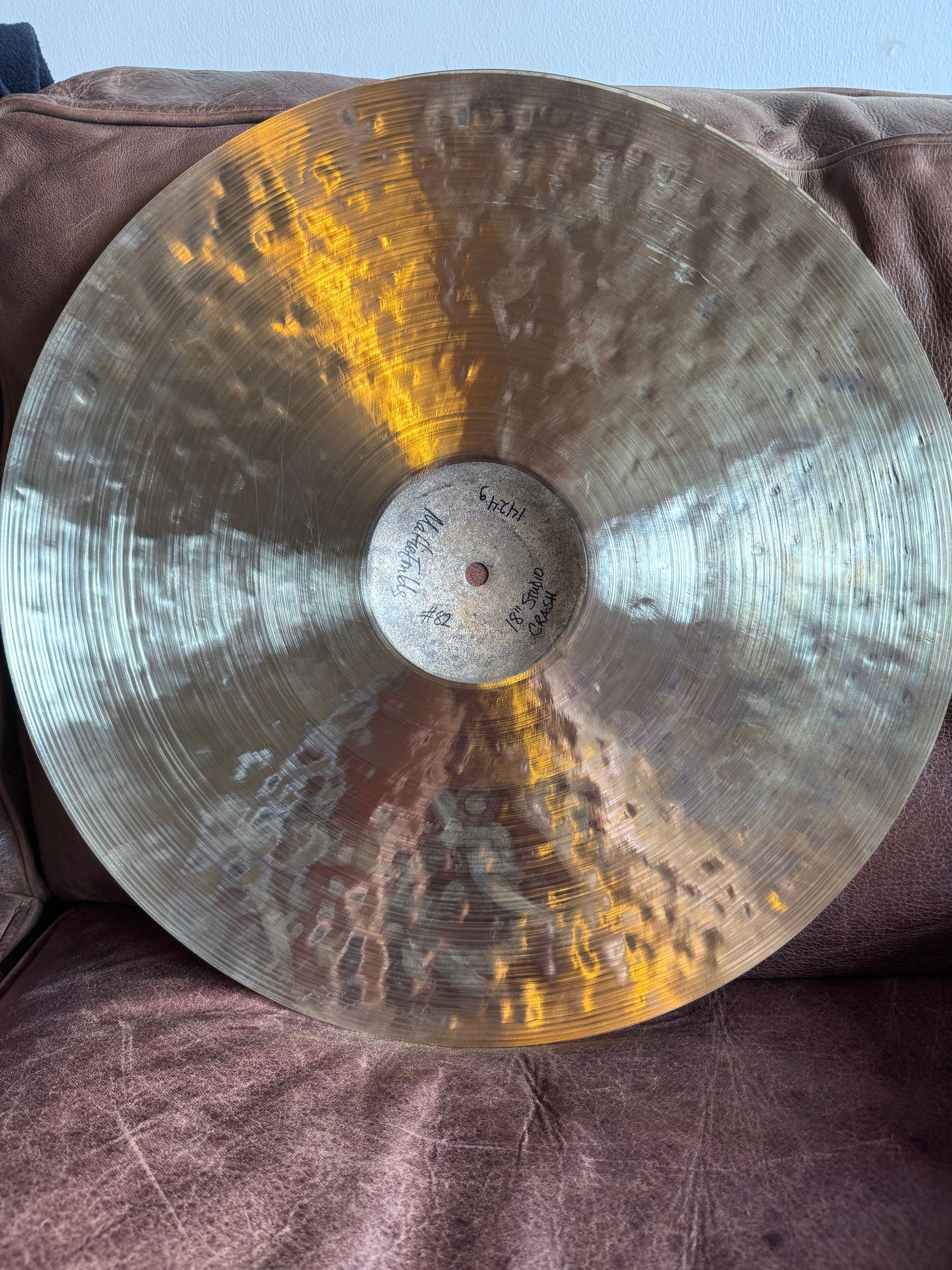 A tarnished, round bronze cymbal lies on a textured, brown surface. It has a central hole.