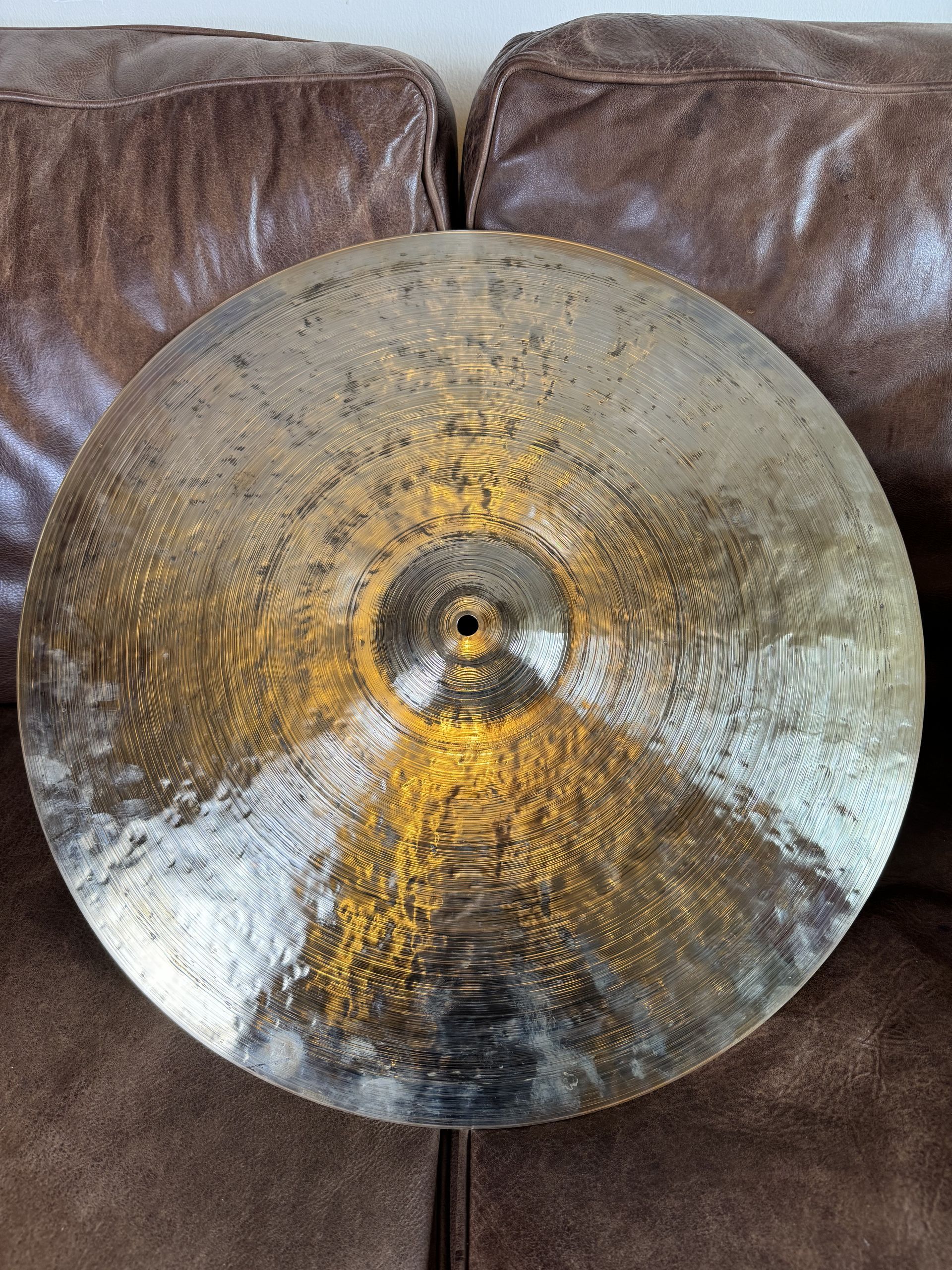 Large round hammered metal cymbal with a gold-silver sheen, leaning against a brown leather couch.