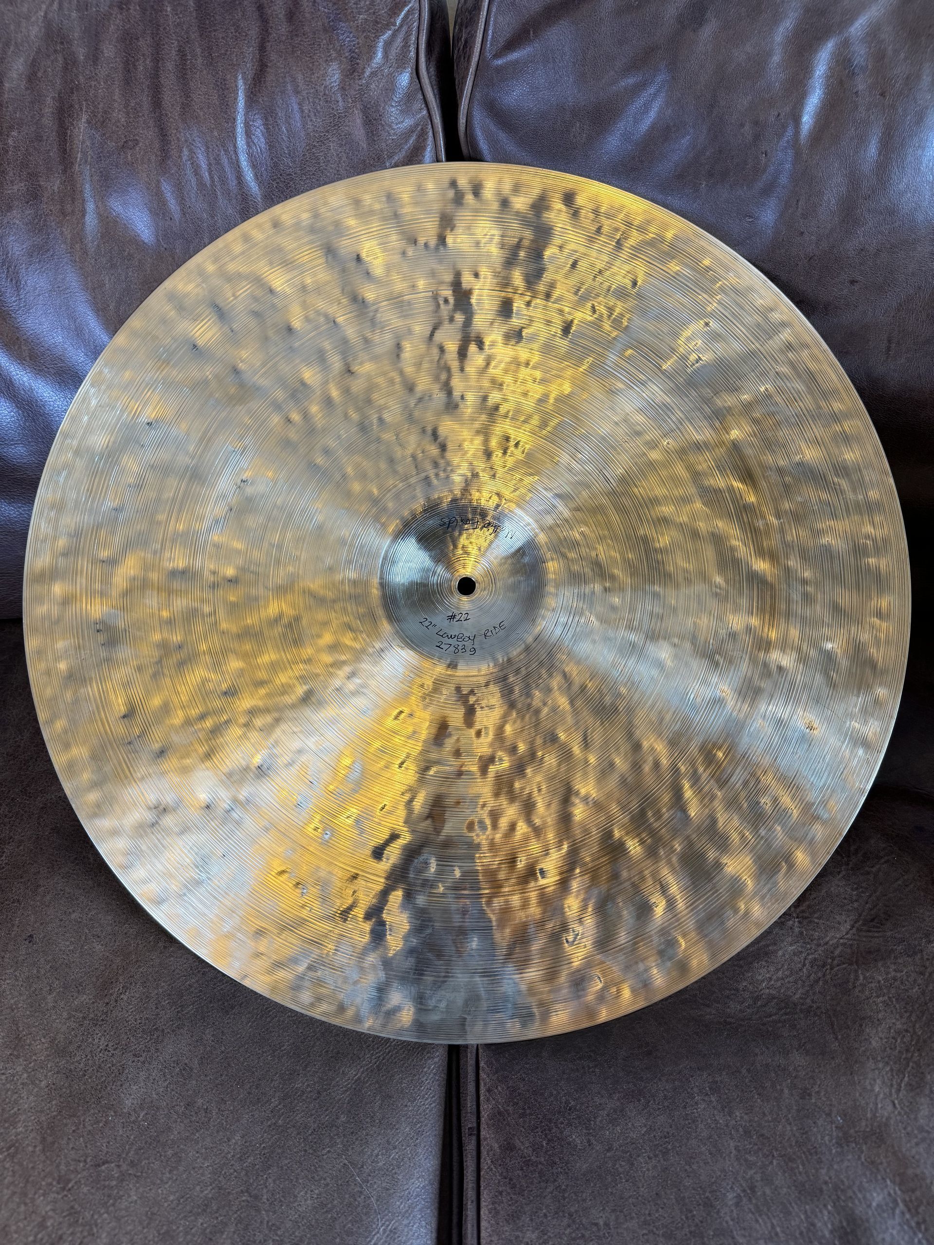 Large round hammered metal cymbal with a gold-silver sheen, leaning against a brown leather couch.