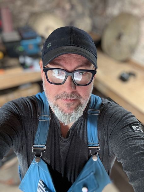 Selfie of a man in a black cap and glasses wearing blue overalls in a workshop.