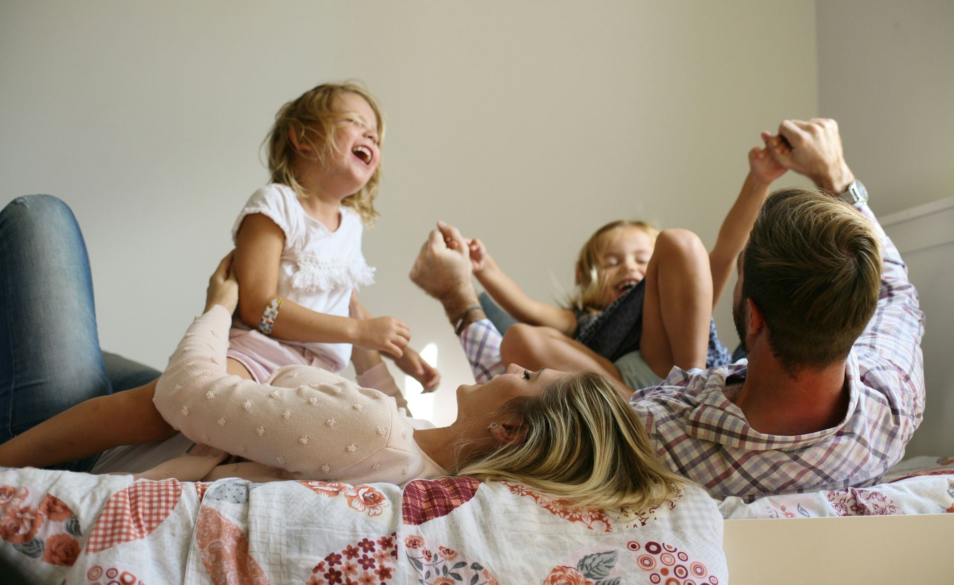 Family playfully interacting on a bed; two children laughing, parents smiling; natural light.