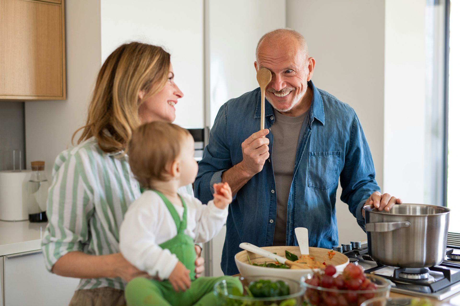 Family in a kitchen, man with wooden spoon to face, woman holding a baby, cooking.