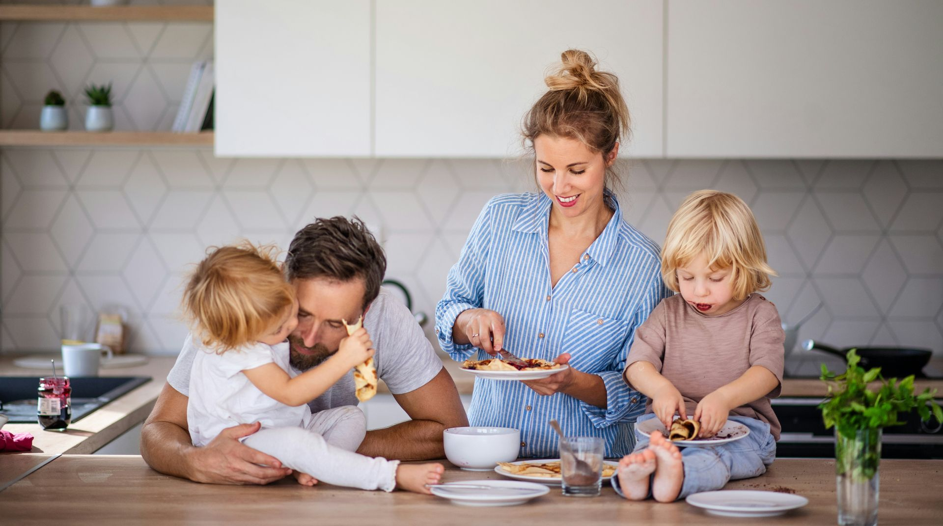 Family in kitchen: Mom cutting pancakes, kids seated on counter, dad watches.